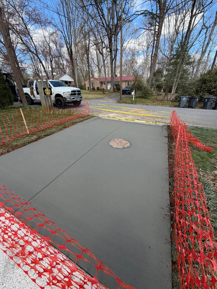 A newly poured concrete driveway with a manhole cover, surrounded by red safety netting and yellow caution tape.